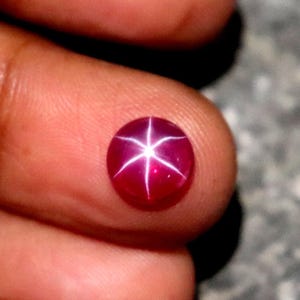 Op de afbeelding: Een ronde, dieprode edelsteen met een zespuntig sterpatroon. De edelsteen wordt tussen vingers gehouden, wat het gepolijste oppervlak en het interne lichtspel benadrukt. De tekst "WestonGems" is zichtbaar.