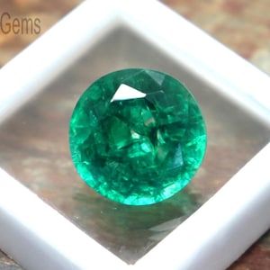 May include: A round, faceted emerald gemstone, displayed in a white square case. The gemstone is a vibrant green color and reflects light. The case is open, revealing the gem's brilliance. The image is set against a natural stone background.