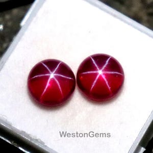 May include: Two round, deep red gemstones with a star-like pattern, displayed in a white plastic case. The gemstones have a polished surface and are set against a white background. The text "WestonGems" is visible on the case.