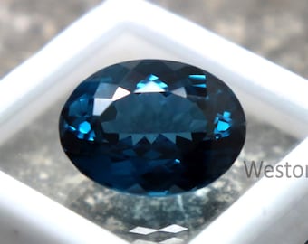 Puede incluir: Una gema ovalada de color verde azulado oscuro, presentada en una caja de plástico blanca cuadrada. La gema tiene múltiples facetas que reflejan la luz, creando un efecto brillante. La gema se encuentra sobre un fondo neutro.
