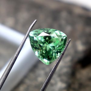 Puede incluir: Una gema verde brillante, facetada y en forma de corazón, es sostenida por unas pinzas. La gema se muestra sobre un fondo borroso, con una caja cuadrada blanca y el texto "WestonGems" visible.