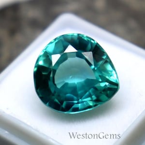 May include: A faceted, teal-colored gemstone, pear-shaped, rests in a white plastic case. The gem has multiple facets that reflect light, creating a sparkling effect. The case is on a dark, textured surface. The text "WestonGems" is visible on the case.