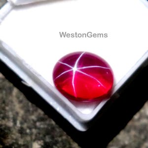 Puede incluir: Una gema roja vibrante con un patrón de estrella, exhibida en una caja de joyería blanca abierta. La gema ovalada refleja la luz, mostrando una estrella de seis rayos. La caja se encuentra sobre un fondo oscuro y texturizado. El texto "WestonGems" es visible.