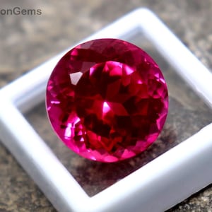 May include: A faceted, round, bright pink gemstone is displayed in a white plastic case. The gem is set against a rough, gray stone surface. The text "WestonGems" is visible on the stone.