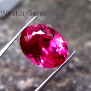 May include: A faceted, oval-shaped, vibrant pink gemstone is held by silver tweezers against a textured gray background. The gem's multiple facets reflect light, creating a sparkling effect. The text "WestonGems" is visible in the background.