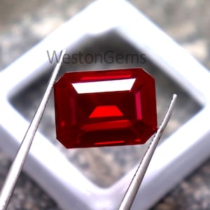 May include: A vibrant, rectangular-cut, red gemstone held by tweezers. The gem is set in a white square frame, with the text "WestonGems" visible. The stone's facets reflect light, showcasing its clarity and rich color. The background is a textured gray surface.