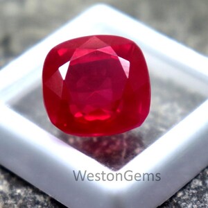 Puede incluir: Una gema roja brillante con talla cojín, presentada en una caja de plástico blanco. La gema tiene múltiples facetas que reflejan la luz, creando un efecto brillante. El fondo es una superficie gris texturizada. El texto "WestonGems" es visible.