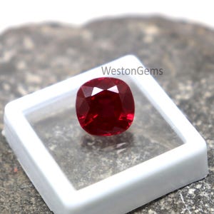 May include: A vibrant, cushion-cut ruby gemstone, presented in a white plastic case. The gem's faceted surface catches the light, producing a bright sparkle. The text "WestonGems" is visible on the top right.