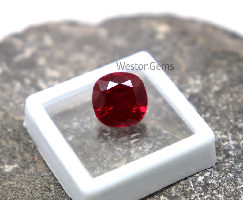 May include: A vibrant, cushion-cut ruby gemstone, presented in a white plastic case. The gem's faceted surface catches the light, producing a bright sparkle. The text "WestonGems" is visible on the top right.
