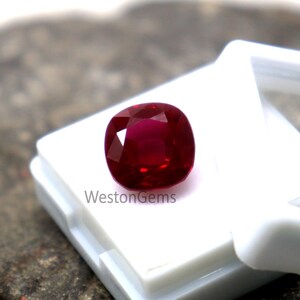 May include: A vibrant, cushion-cut ruby gemstone, deep red in colour, presented in a white plastic case. The polished gemstone is set against a neutral backdrop. The text "WestonGems" is visible on the case.