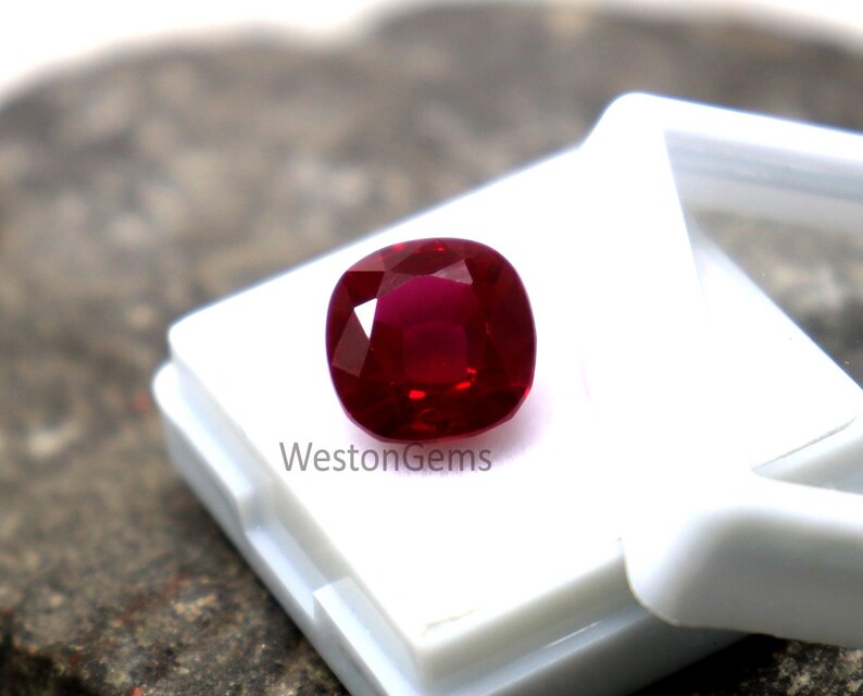 May include: A vibrant, cushion-cut ruby gemstone, deep red in colour, presented in a white plastic case. The polished gemstone is set against a neutral backdrop. The text "WestonGems" is visible on the case.