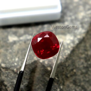May include: A faceted, cushion-cut ruby gemstone held by tweezers. The deep red gem is displayed against a textured, grey surface. The white container in the background is out of focus. The text "WestonGems" is visible.