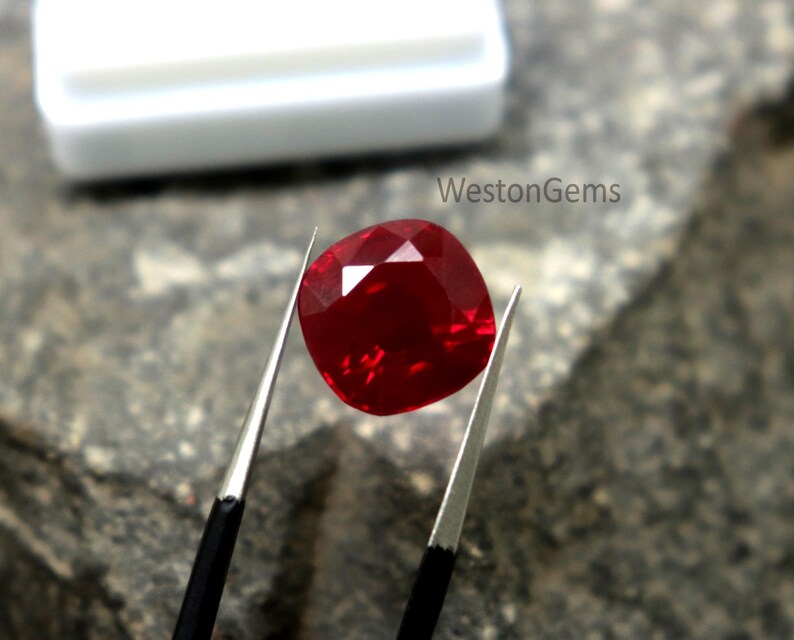 May include: A faceted, cushion-cut ruby gemstone held by tweezers. The deep red gem is displayed against a textured, grey surface. The white container in the background is out of focus. The text "WestonGems" is visible.