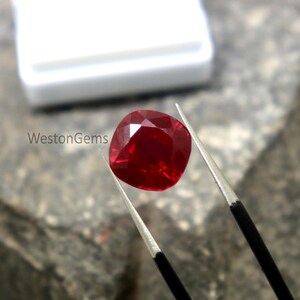 May include: A faceted, cushion-cut ruby gemstone held by tweezers. The ruby is a deep, vibrant red colour and reflects light. The image includes the text "WestonGems".