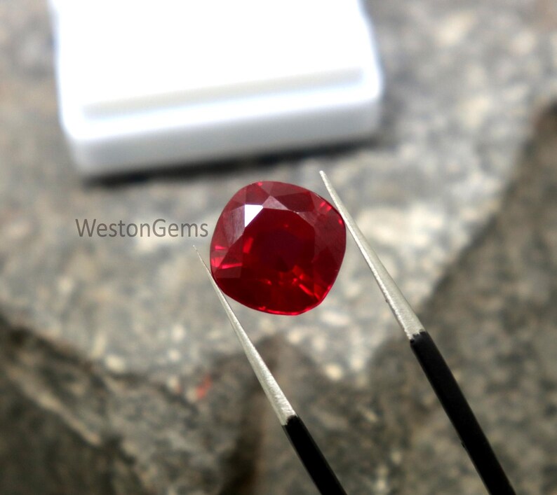 May include: A faceted, cushion-cut ruby gemstone held by tweezers. The ruby is a deep, vibrant red colour and reflects light. The image includes the text "WestonGems".