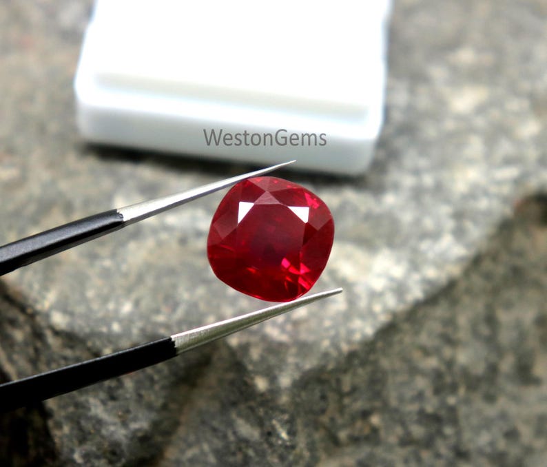 May include: A vivid red cushion-cut gemstone is held by tweezers against a textured surface. The gemstone has a faceted surface that reflects light, showcasing its brilliance. The white container in the background has the text "WestonGems".