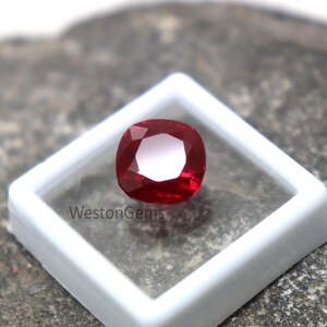 May include: A vibrant red, cushion-cut gemstone, presented in a white plastic display case. The gem's facets catch the light, creating a sparkling effect. The text "WestonGems" is printed on the case.