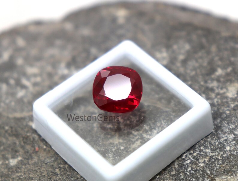 May include: A vibrant red, cushion-cut gemstone, presented in a white plastic display case. The gem's facets catch the light, creating a sparkling effect. The text "WestonGems" is printed on the case.