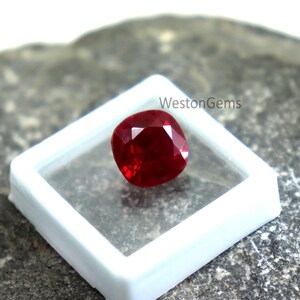 May include: A vivid red gemstone, cushion-cut, presented in a clear, square container. The gem reflects light, showcasing its brilliance. The container sits on a textured, grey surface. The text "WestonGems" is visible.