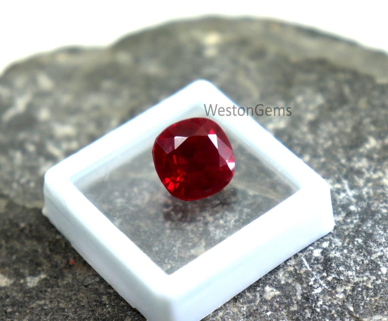 May include: A vivid red gemstone, cushion-cut, presented in a clear, square container. The gem reflects light, showcasing its brilliance. The container sits on a textured, grey surface. The text "WestonGems" is visible.