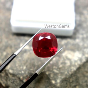 May include: A deep red cushion-cut ruby gemstone, held by tweezers. The ruby reflects light, highlighting its facets. The text "WestonGems" is visible in the background.