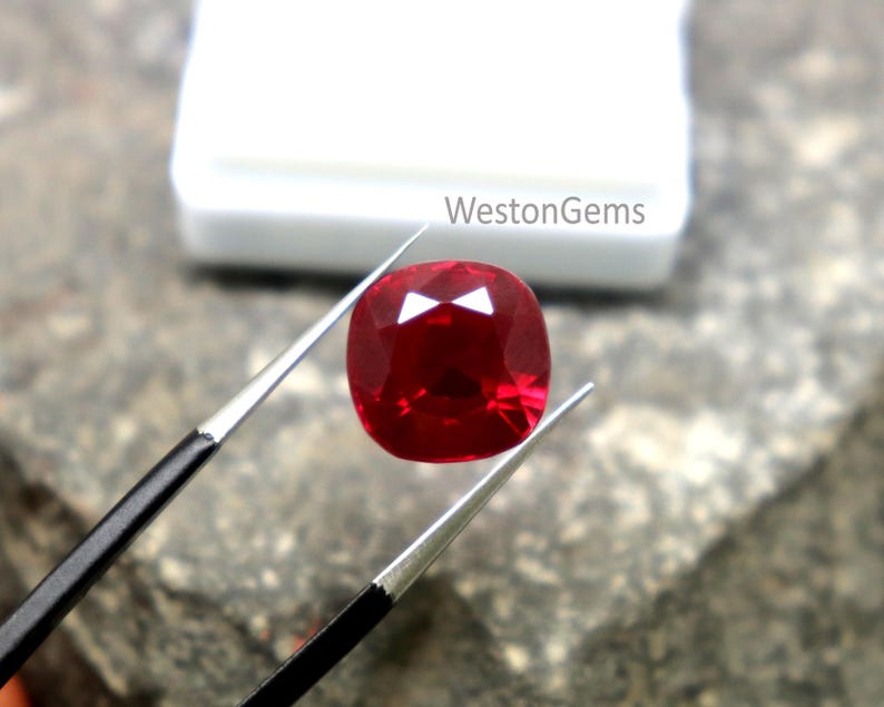 May include: A deep red cushion-cut ruby gemstone, held by tweezers. The ruby reflects light, highlighting its facets. The text "WestonGems" is visible in the background.