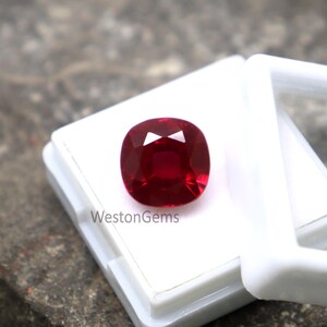 May include: A cushion-cut ruby gemstone, displayed in a white case. The ruby is a rich, deep red, with multiple facets that reflect light. The white case provides a clean backdrop, accentuating the gemstone's brilliance. The text "WestonGems" is visible on the case.