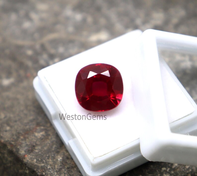 May include: A cushion-cut ruby gemstone, displayed in a white case. The ruby is a rich, deep red, with multiple facets that reflect light. The white case provides a clean backdrop, accentuating the gemstone's brilliance. The text "WestonGems" is visible on the case.