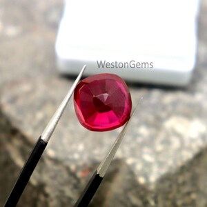 May include: A cushion-cut ruby gemstone, held by tweezers. The ruby is a deep red colour and has multiple facets. The text "WestonGems" is visible in the background.