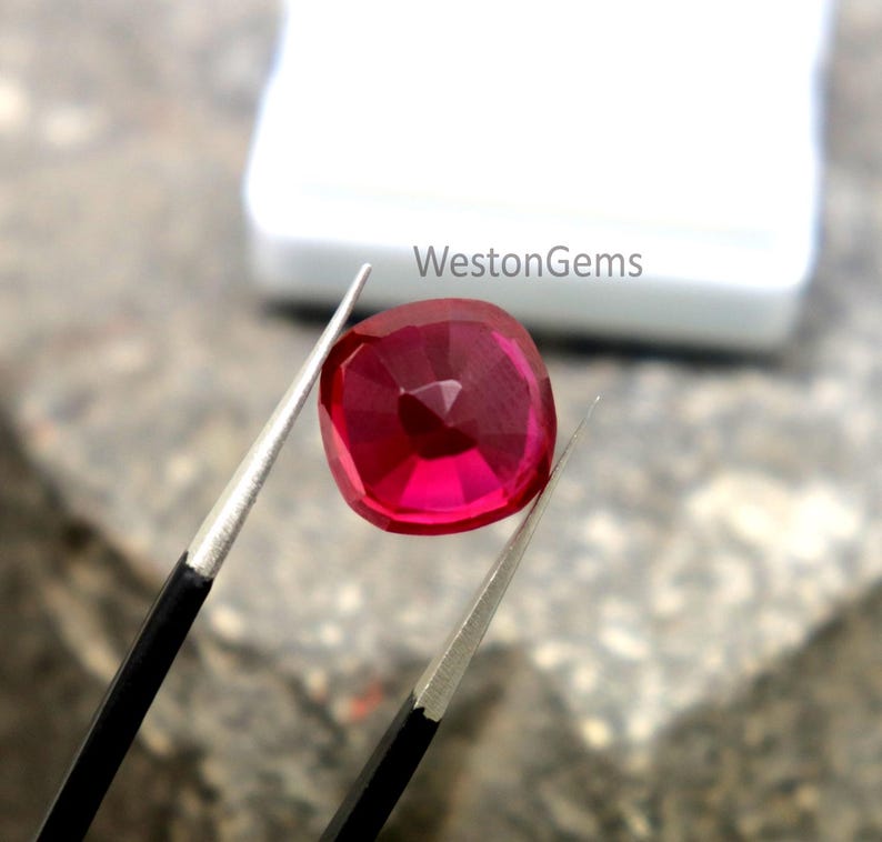 May include: A cushion-cut ruby gemstone, held by tweezers. The ruby is a deep red colour and has multiple facets. The text "WestonGems" is visible in the background.