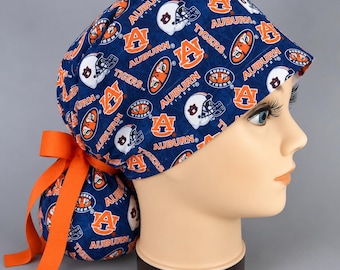 Può includere: Una cuffia chirurgica blu navy con una stampa all-over del logo Auburn Tigers, caschi da football americano e la scritta Auburn in arancione e bianco. La cuffia presenta una parte posteriore arricciata fissata con un nastro arancione vivo.