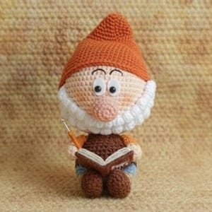 Peut inclure: Une poupée gnome au crochet avec un chapeau orange, une barbe blanche et un corps marron. Le gnome tient un livre et un pointeur. La poupée a de grands yeux globuleux et porte un pantalon bleu. Le fond est une surface texturée et beige.