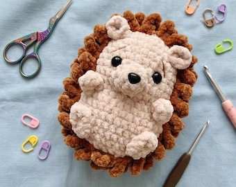 May include: A handmade crochet hedgehog in light brown and tan yarn. The hedgehog has a round body, small ears, and black safety eyes. Crochet hooks, stitch markers, and rainbow scissors are also in the image.