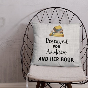 May include: A white throw pillow with a black and white graphic of a stack of books. The text on the pillow reads "Reserved for Andrea and her book".