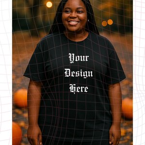 以下が含まれることがあります： ゴシックフォントで「Your Design Here」と書かれた黒いTシャツ。その人は笑顔でTシャツを着ています。背景には、カボチャとボケライトのあるぼやけた秋のテーマがあります。