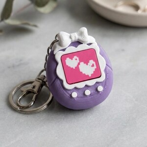 Op de afbeelding: Een paarse sleutelhanger met een retro handheld game ontwerp. De sleutelhanger heeft een witte strik detail aan de bovenkant en een roze scherm met twee witte pixelhartjes. De sleutelhanger is bevestigd aan een zilveren sleutelring.