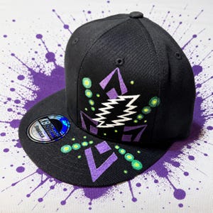 Puede incluir: Gorra snapback negra con visera plana, con un diseño geométrico morado y blanco con detalles verdes. La gorra tiene un botón redondo en la parte superior y una pegatina en la visera.