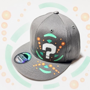 May include: A grey snapback cap featuring a white question mark graphic on the front panel, accented with mint green curved lines and small orange circular dots. A circular holographic sticker with the text KBETHOS is on the flat brim.
