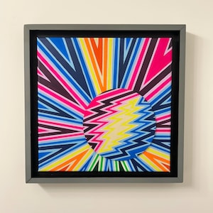 May include: A framed canvas print with a colorful, abstract design. The design features a circle with a lightning bolt pattern in the center, surrounded by radiating lines of various colors.