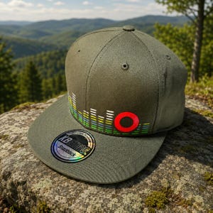 Può includere: Cappellino snapback verde oliva con visiera piatta. Il cappellino presenta una grafica di onde sonore colorate e un cerchio rosso. La visiera ha un adesivo olografico con la scritta "KBETHOS Snapback".