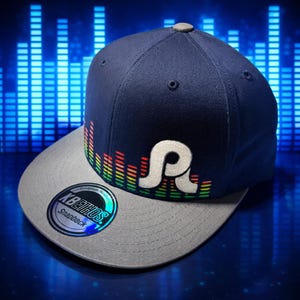 Puede incluir: Gorra snapback azul marino con visera gris. La gorra presenta un logotipo "R" blanco estilizado y un diseño de ecualizador colorido. La gorra tiene una pegatina "KBETHOS Snapback" en la visera.