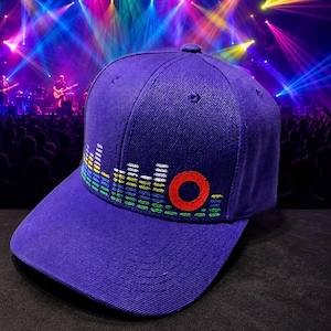 May include: A purple baseball cap featuring an embroidered graphic of a multi-colored sound equalizer bar and a prominent red circle on the front panel. The cap has a structured crown, a curved brim, and stitched eyelets on the top panels.