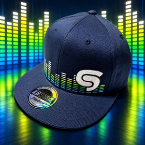 May include: A navy blue snapback cap featuring an embroidered white letter S and a colorful sound wave graphic. A circular sticker on the flat brim displays the text KBETHOS Premium Headwear Snapback in a holographic font against a black background.