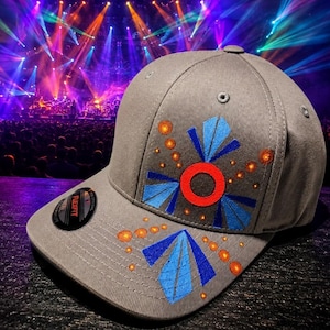 May include: A grey baseball cap featuring a geometric design on the front panel and brim. The pattern consists of a central red circle, blue triangular shapes, and small orange dots. A black circular sticker with the text Flexfit is on the brim.