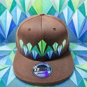 May include: A brown snapback cap featuring a geometric pattern of green and blue triangles across the front panels. The flat brim displays a circular sticker with the text KBETHOS Snapback. The cap has a structured crown and stitched eyelets.