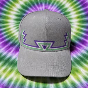 Puede incluir: Gorra de béisbol gris con un diseño geométrico en morado y verde. La gorra tiene una visera curva y una corona estructurada. El diseño es una serie de zigzags y triángulos. El fondo es un patrón tie-dye en tonos verdes, morados y blancos.