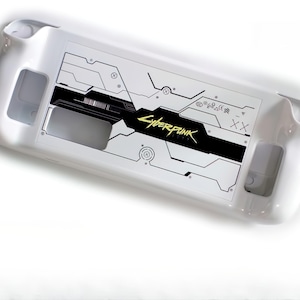 Puede incluir: Funda blanca para consola de juegos con un diseño Cyberpunk negro y amarillo. La funda presenta un patrón geométrico y el logotipo del juego. La funda tiene recortes para botones y puertos.