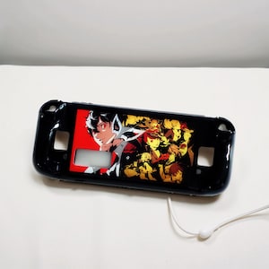 May include: Black protective case for a handheld gaming device. The case features a colourful graphic of anime characters with a red and yellow colour scheme. The case has cutouts for buttons and ports.