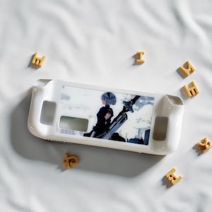 Könnte beinhalten: Eine weiße Schutzhülle für ein Handheld-Gaming-Gerät mit einem Grafikmotiv eines Charakters mit blauen Haaren und einem Schwert. Buchstabenförmige Cracker umgeben die Hülle auf einer weißen Oberfläche.