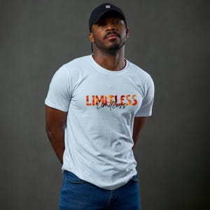 Puede incluir: Camiseta blanca con la palabra "LIMITLESS" en letras de bloque naranja y rojo, con la palabra "limitless" en cursiva debajo. La persona de la imagen lleva una gorra de béisbol negra y vaqueros azules.